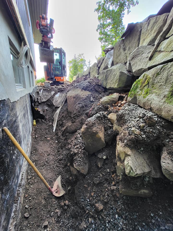 Grøft gravd for drenering langs grunnmur med gravemaskin i bakgrunnen